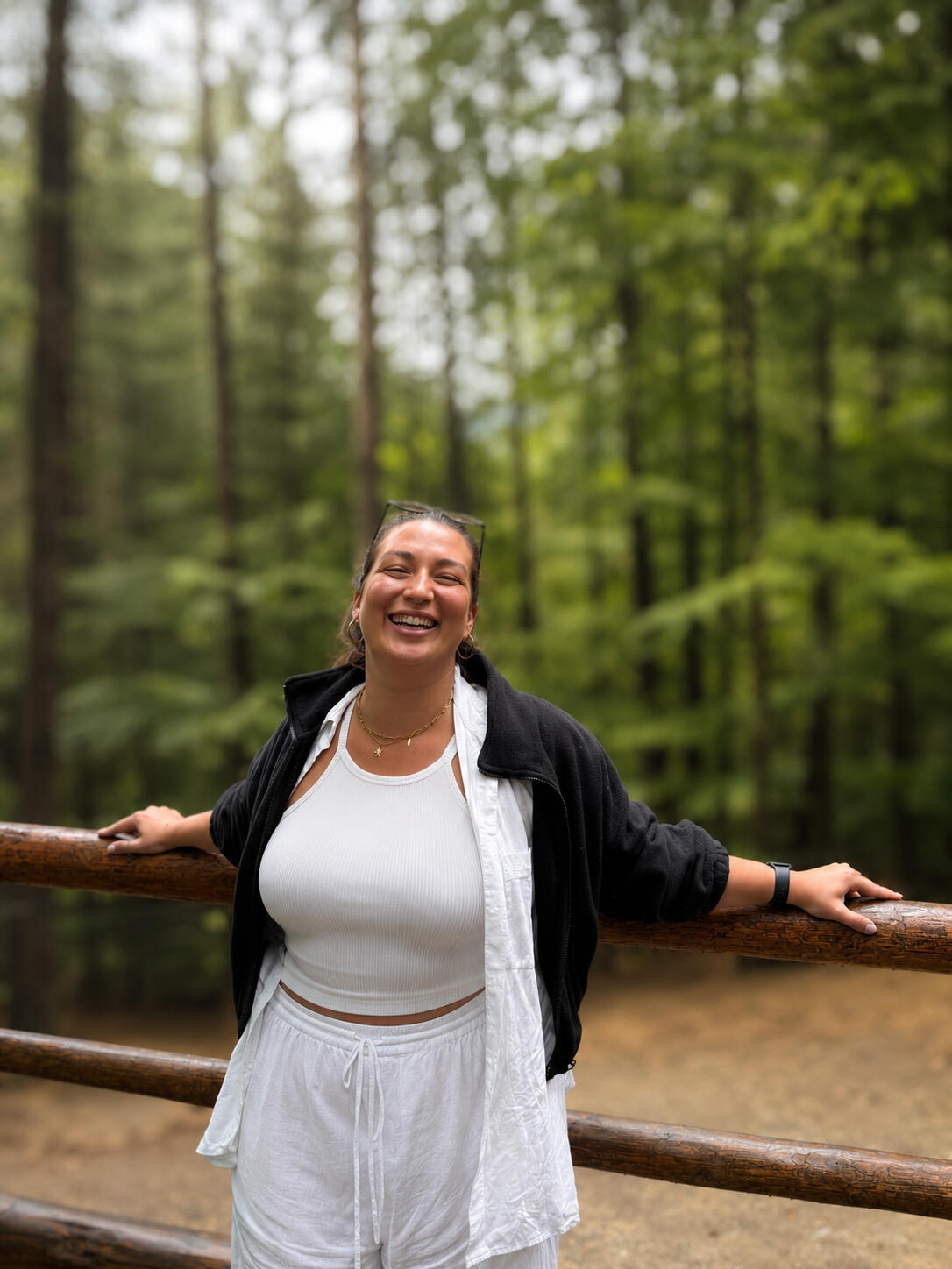Jaz is standing in front of a bright forest, with her arms open, resting on a wooden fence, she is smiling/laughing.